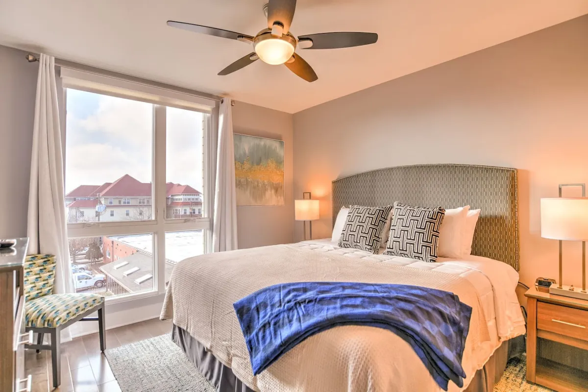 A serene king bedroom featuring a comfortable bed adorned with patterned pillows, a window that fills the room with natural light, and a tasteful decorative chair. Subtle, modern decor creates a restful and welcoming ambiance, ideal for relaxation or enjoying a quiet morning.