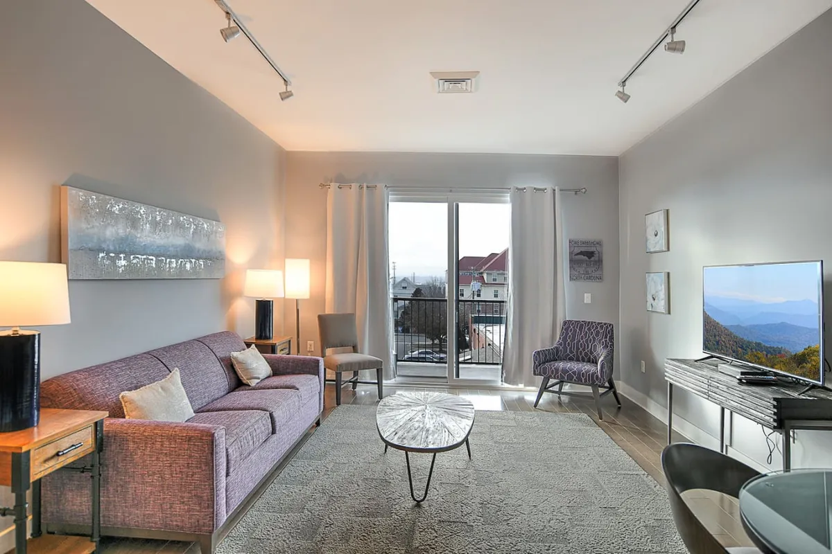 The image features a cozy living area with a sofa, coffee table, TV, and a visible balcony view through glass doors. The space is arranged for intimacy—ideal for gathering with friends, watching TV, or enjoying the sunset from the balcony.