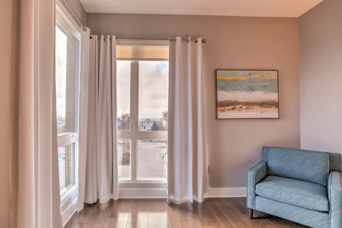 A spacious, light-filled living area featuring large windows, a comfortable blue accent chair, and an abstract painting. The scene captures subtle details like streaming natural light and an inviting atmosphere perfect for relaxing or connecting with friends and family.