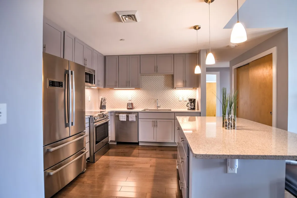 A warm, modern kitchen scene featuring granite countertops, stainless steel appliances, under-cabinet lighting, and pendant lights. The layout is inviting for hosting or enjoying casual meals, accentuated by morning sunlight streaming in, which adds a cozy, homely feel.
