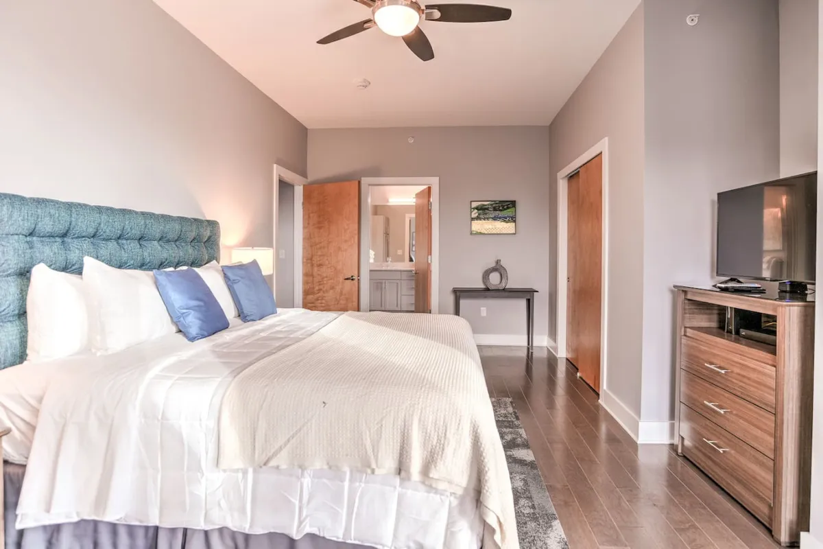 A second king bedroom with blue accent pillows, a wooden dresser, TV stand, and curated artwork. The space feels both stylish and cozy, offering a relaxing environment for reading, watching TV, or unwinding after a day out. Natural light pours in from a window, enhancing the soothing ambiance.