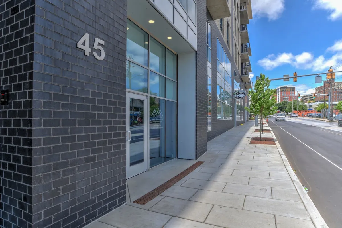 This image captures the building's modern exterior facade, with a clear view of the entrance door, address number, sidewalk, and surrounding urban environment. The bustling sidewalk and visible traffic lights emphasize the downtown location and easy access to the neighborhood's amenities.