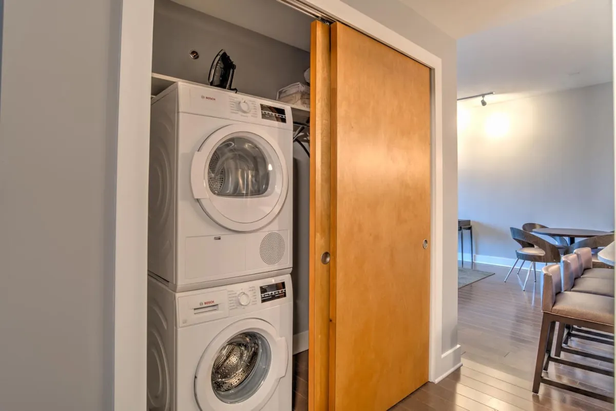 A functional and inviting view of the entryway area, highlighting a stacked washer and dryer set behind a wooden sliding door, with a glimpse of a dining area in the background. The design is both practical and cozy, reflecting attention to convenience in daily living.