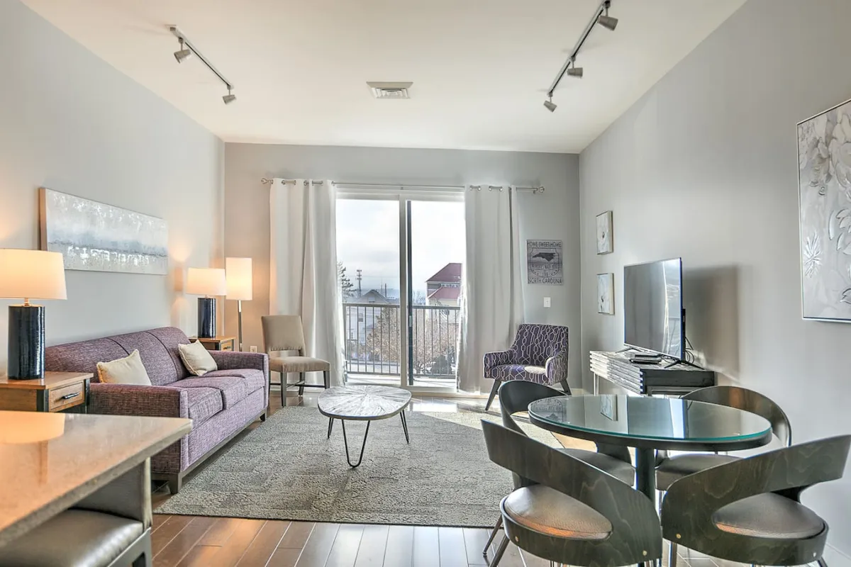 This image showcases a comfortable living room with a sofa, coffee table, large windows, and a television. Morning light streams through the expansive windows, creating a bright and inviting atmosphere. The seating is plush and arranged for both relaxation and conversation, with views out over the city.