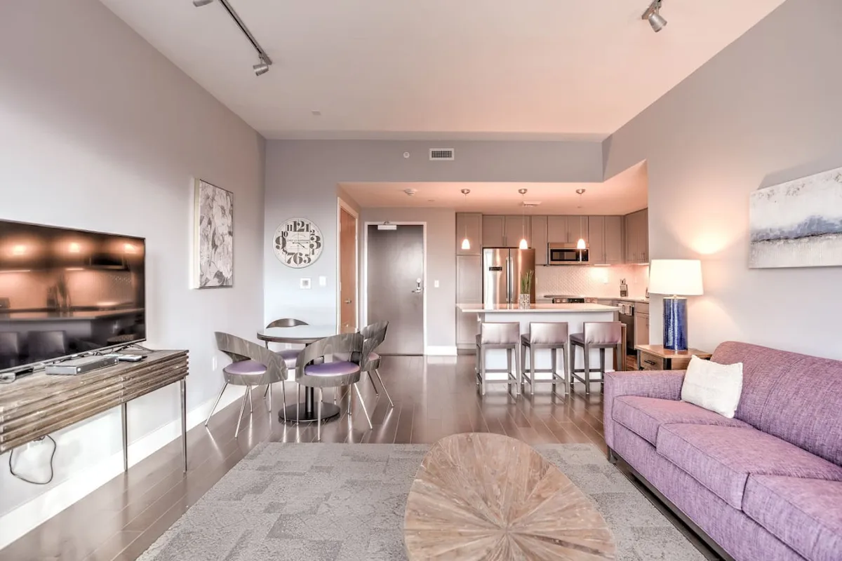 The open-concept space includes a dining table and kitchen island, with a purple sofa and television in the background. The scene emphasizes opportunities for casual dining, socializing, and relaxing, illustrating how the kitchen and dining nook integrate with living areas.
