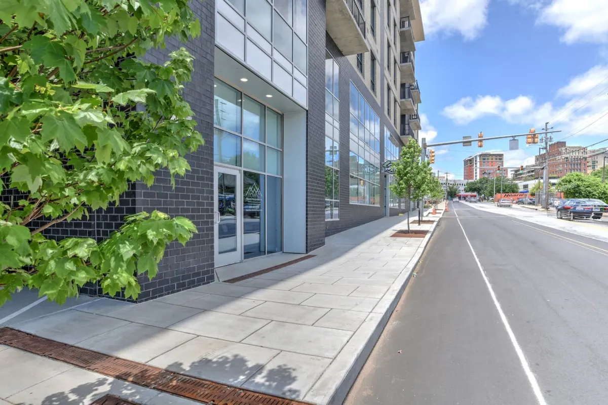 A dynamic streetscape featuring the exterior of a modern building with expansive glass windows, sleek architectural lines, and mature street trees. The sidewalk is bustling with people enjoying the sunny day, suggesting easy access to shops, cafes, and local attractions.